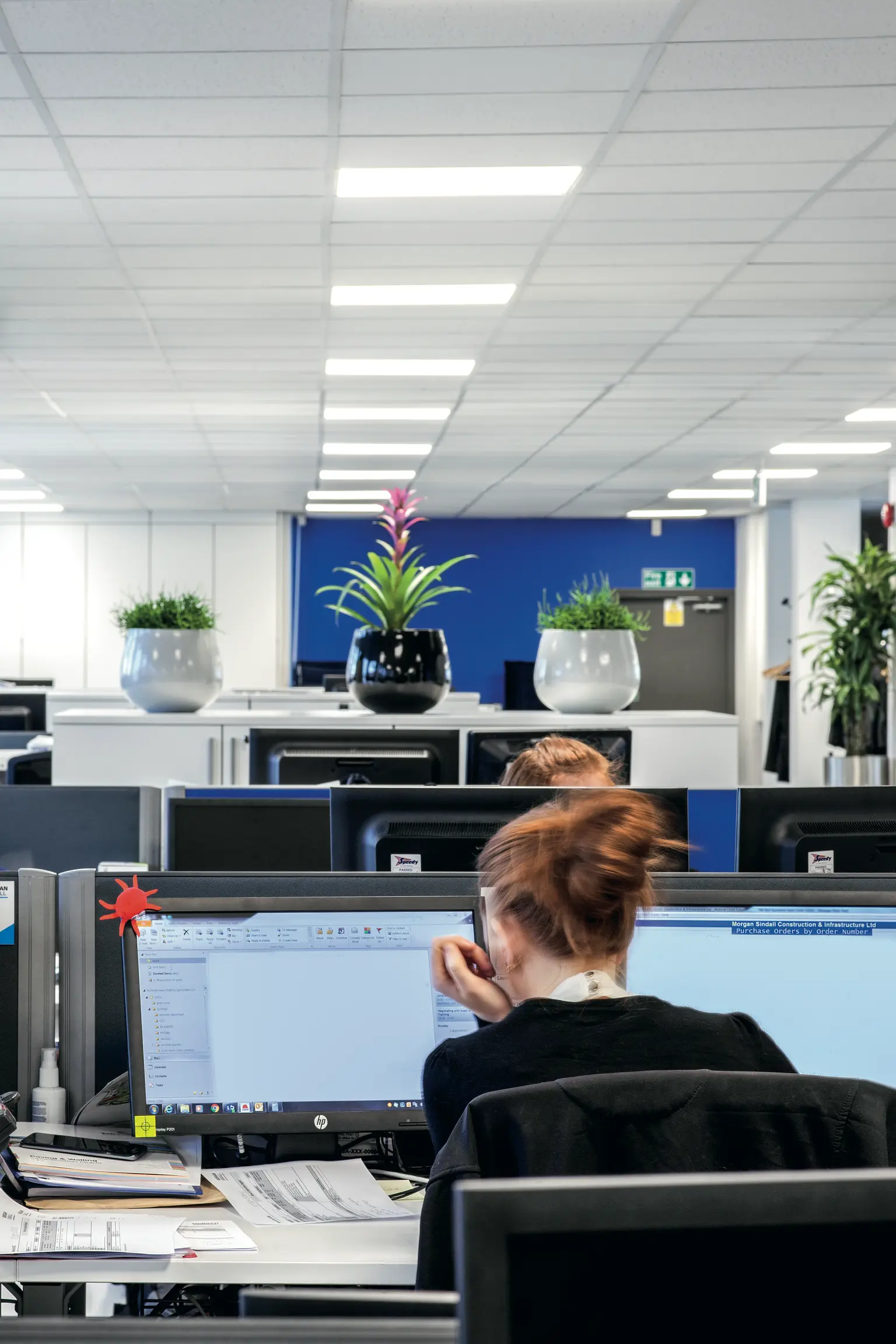 Ein modernes Büro mit einer Besprechungsraum mit Glaswänden neben einem offenen Arbeitsbereich mit Computern und Bürostühlen. Das Raumkonzept ist offen und luftig, mit viel natürlichem und künstlichem Licht.