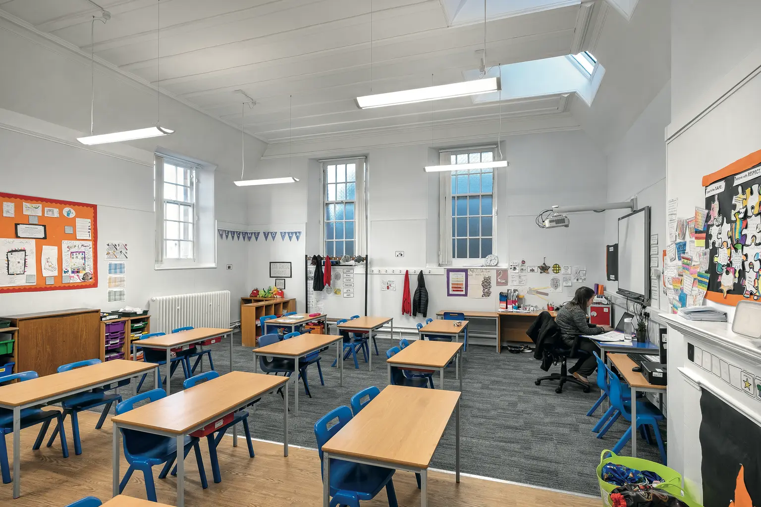 Un’aula luminosa con banchi in legno, sedie azzurre e grandi finestre che lasciano entrare la luce naturale. Le pareti sono decorate con materiali didattici e lo spazio include una lavagna e luci montate a soffitto.