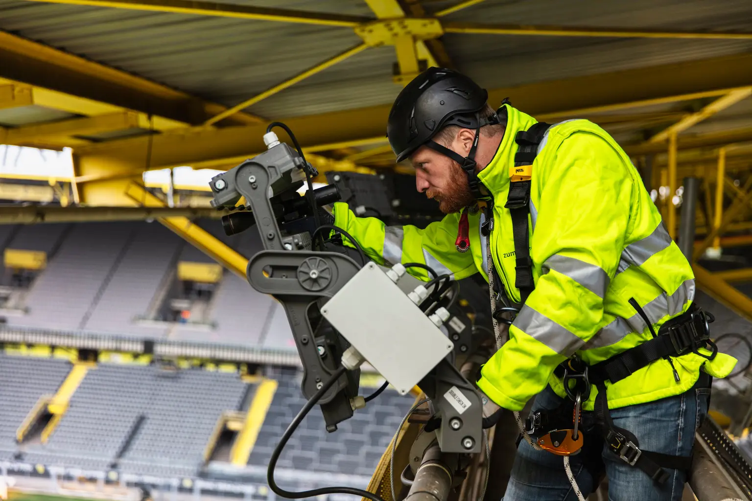 Ein Techniker in einer gut sichtbaren Jacke und Sicherheitsausrüstung arbeitet an Geräten im Signal Iduna Park. Im Hintergrund sind die ikonische gelbe Stahlkonstruktion und die gestuften Sitzreihen des Stadions zu sehen.