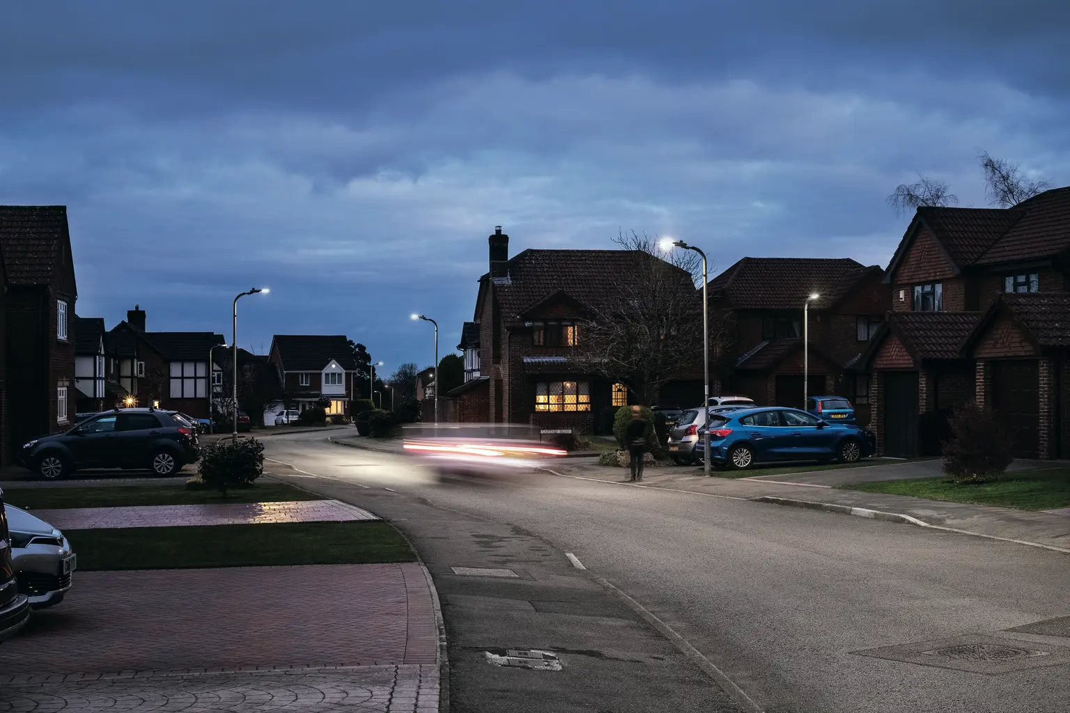 Una tranquilla strada di periferia al crepuscolo con villette e auto parcheggiate su entrambi i lati. I lampioni sono accesi e un'auto passa, lasciando scie di luce sulla strada.