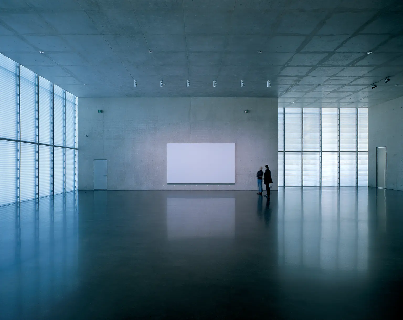 Ein minimalistischer, lichtdurchfluteter Raum im Kunsthaus Bregenz mit glatten Betonwänden, einer großen leeren Leinwand und zwei Personen, die den Raum betrachten. Die klare Architektur betont die moderne Ästhetik des Gebäudes.