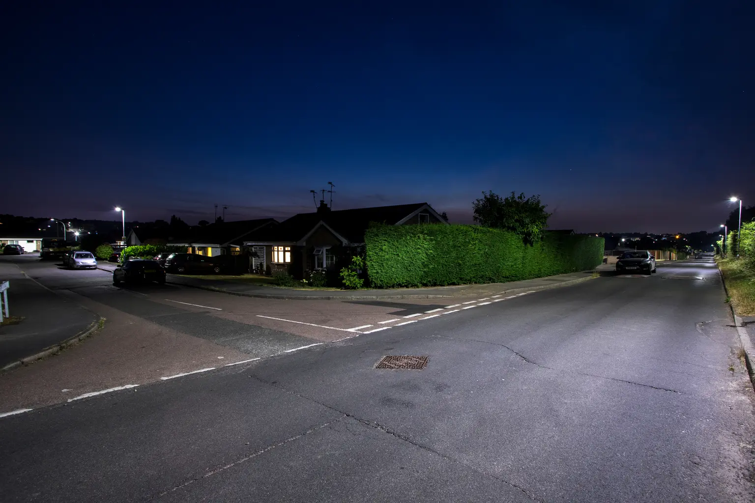 Una tranquilla strada residenziale di notte è illuminata dai lampioni, con case e alte siepi lungo il ciglio della strada. Sono visibili diverse auto parcheggiate e il cielo è di un azzurro profondo.