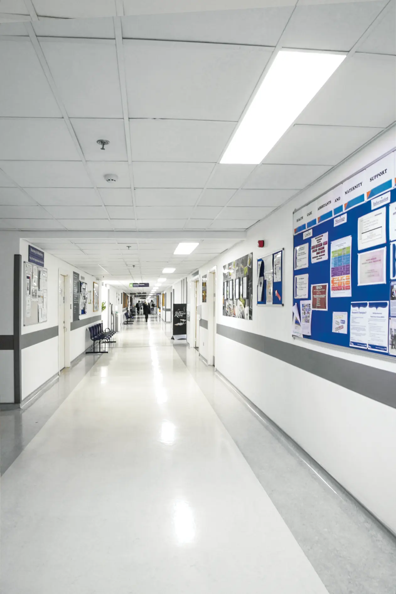 Corridoio ospedaliero luminoso con pareti bianche e dettagli grigi. Bacheche visualizzano gli avvisi. Illuminato da un pannello LED.