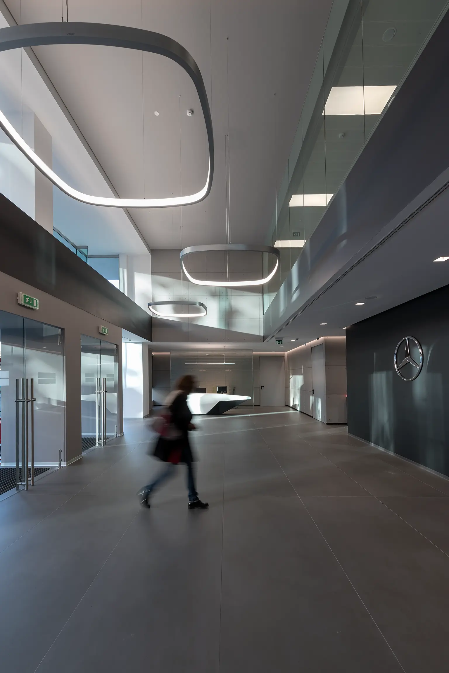 Un moderno atrio d'ufficio con soffitti alti, grandi lampade geometriche e un logo Mercedes-Benz sul muro. Una persona attraversa l'area d'ingresso, ampia e minimalista.