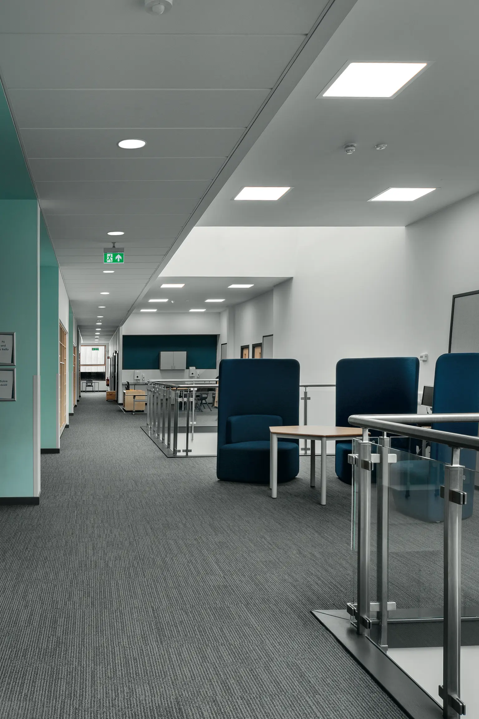 Corridoio scolastico moderno con pavimenti in moquette e vinile, pareti bianche e dettagli color foglia di tè. Lo spazio include panche con tappezzeria color azzurro, tavoli per attività collaborative e apparecchi a soffitto incassati per un'illuminazione intensa e vivace.
