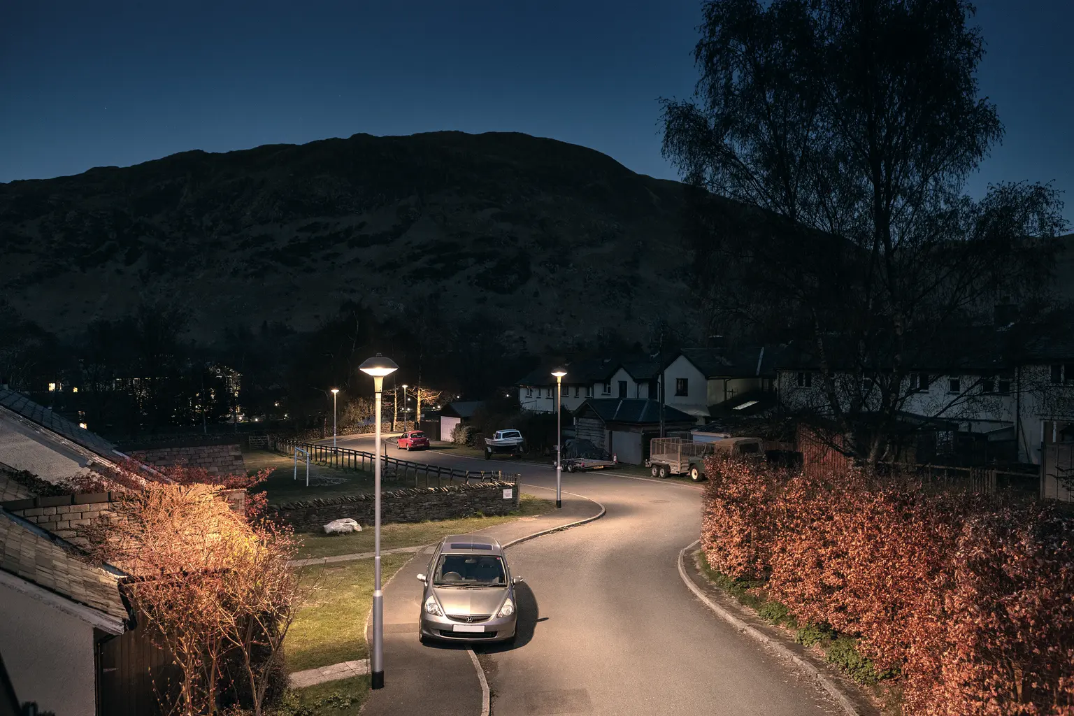 Una tranquilla strada residenziale di notte con una sola auto parcheggiata, illuminata dai lampioni. Case e alberi fiancheggiano la strada, con colline scure sullo sfondo.