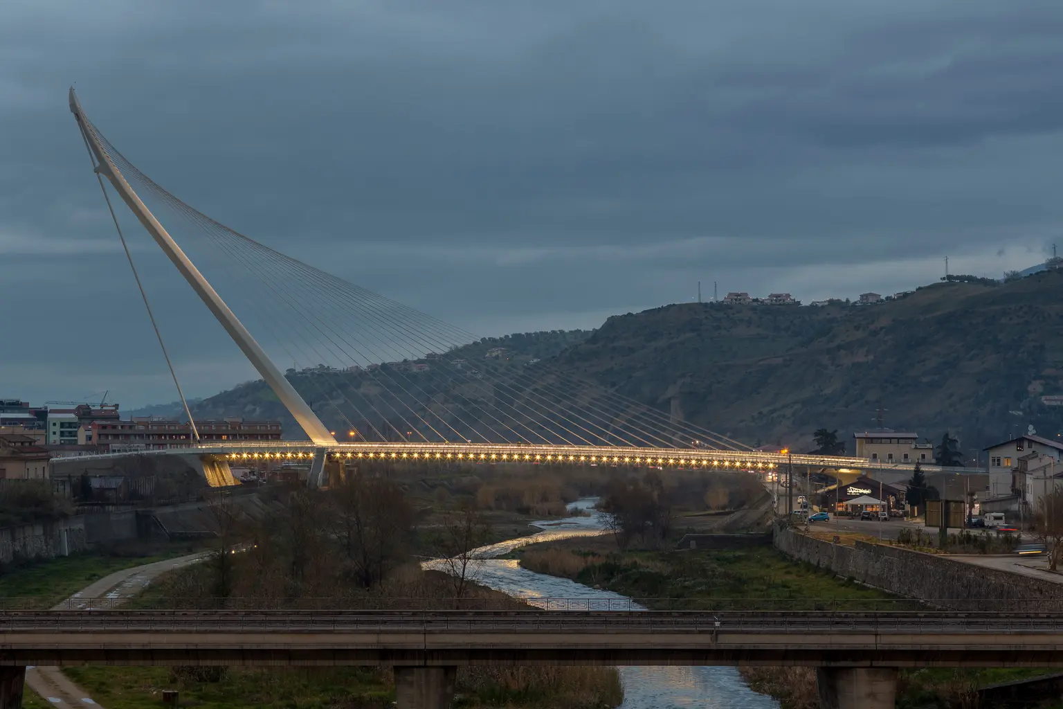 Moderne Schrägseilbrücke bei Dämmerung, beleuchtet und über einen Fluss führend. Im Hintergrund sind bewaldete Hügel und einige Gebäude zu sehen.