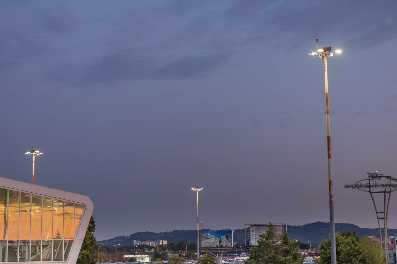 Area esterna al crepuscolo con alti proiettori che illuminano la scena. Sullo sfondo, gli edifici della città e le colline sono visibili sotto un cielo sereno della sera.