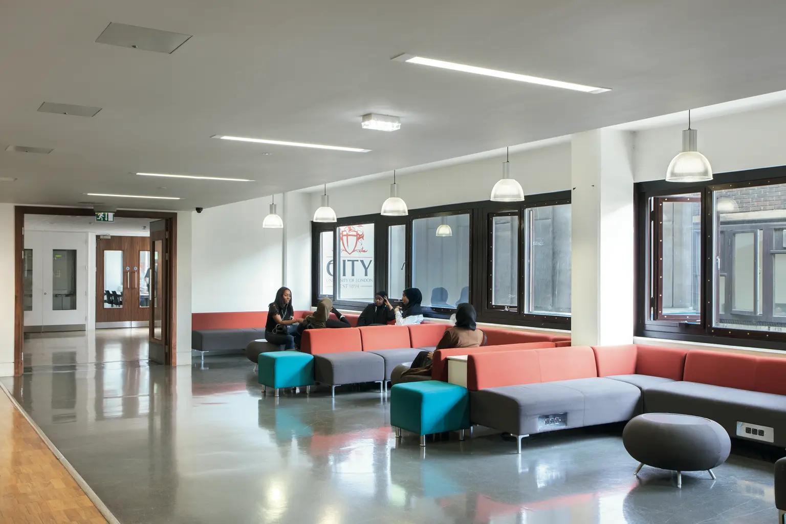 Un'area lounge scolastica moderna con posti a sedere colorati, tra cui divani rossi e divani color foglia di tè e pouf grigi. Lo spazio è ben illuminato da luci a sospensione e incassate, con ampie finestre per la luce naturale.