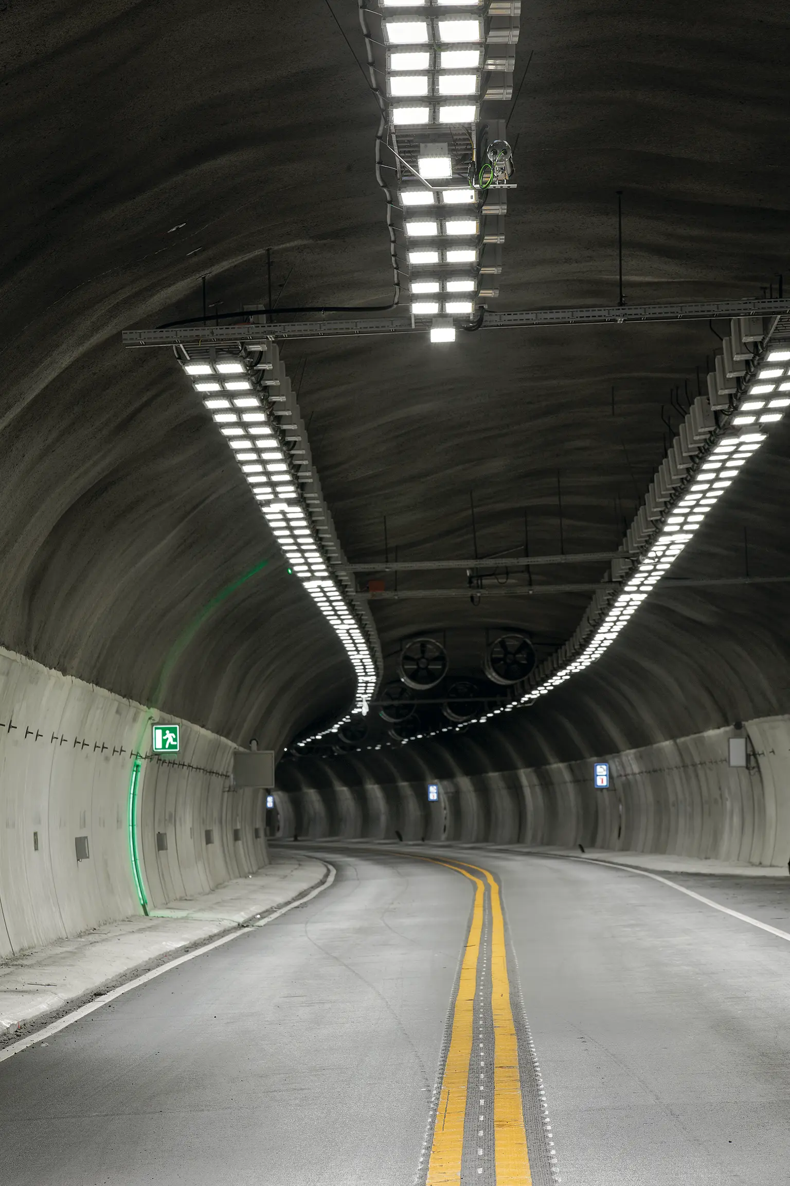 Un tunnel stradale curvo con pareti in cemento liscio, luci LED luminose sul soffitto e un segnale di uscita di emergenza verde brillante sulla sinistra. La strada a due corsie è segnalata da linee gialle e presenta un layout aperto per garantire un flusso di traffico efficiente.