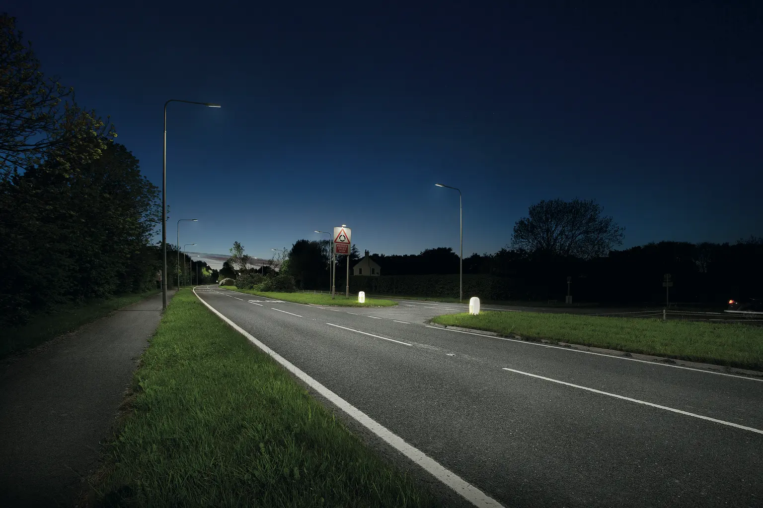 Eine ruhige Vorortstraße bei Nacht, beleuchtet von modernen Straßenlaternen, mit Grasflächen und Bäumen auf beiden Seiten. Der Himmel ist dunkelblau und in der Ferne sind Häuser zu erkennen.