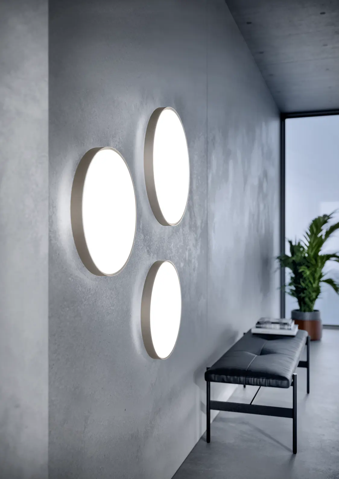 Tre apparecchi da parete a LED LANOS illuminano un corridoio moderno con pareti in cemento e piante, mentre sotto si trova una panchina in stile minimalista.