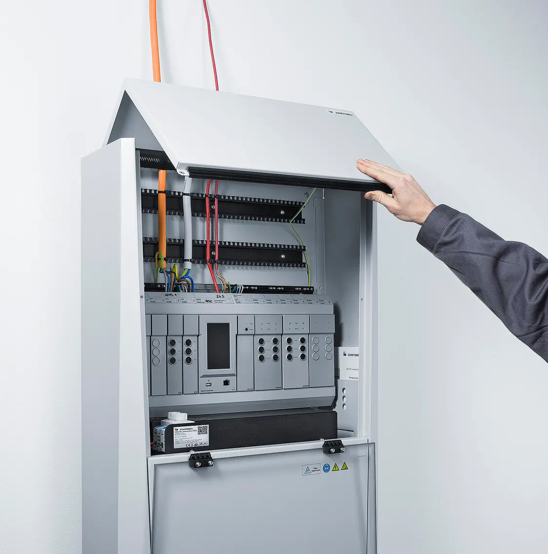  L'armadio del sistema di alimentazione di emergenza ONLITE central eBox viene aperto con una mano, rendendo visibili i cavi e i componenti elettronici all'interno.