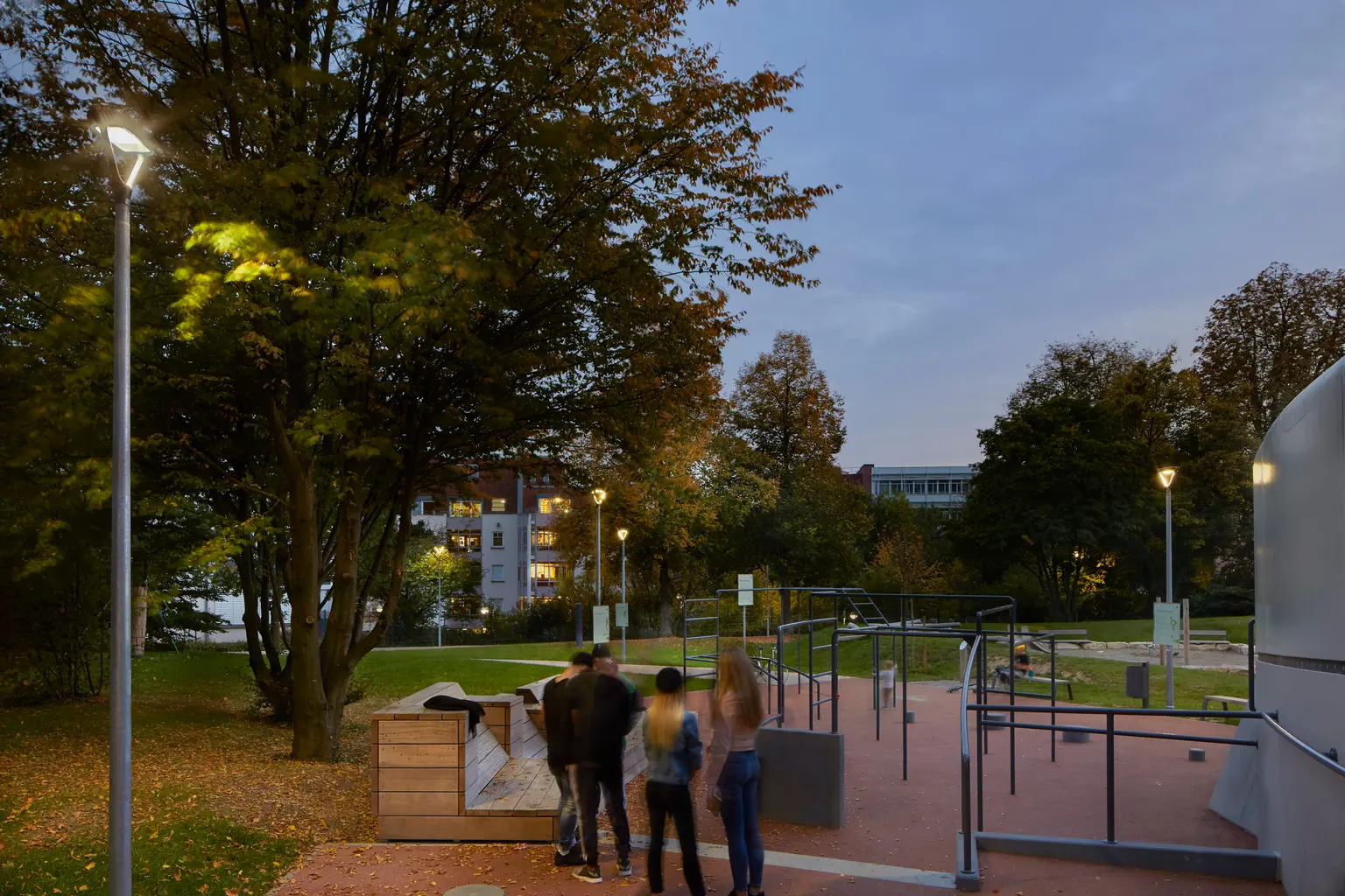 Menschen versammeln sich in der Nähe von Outdoor-Fitnessgeräten in einem Park, wobei die Beleuchtung für gute Sichtbarkeit und eine einladende Atmosphäre sorgt.