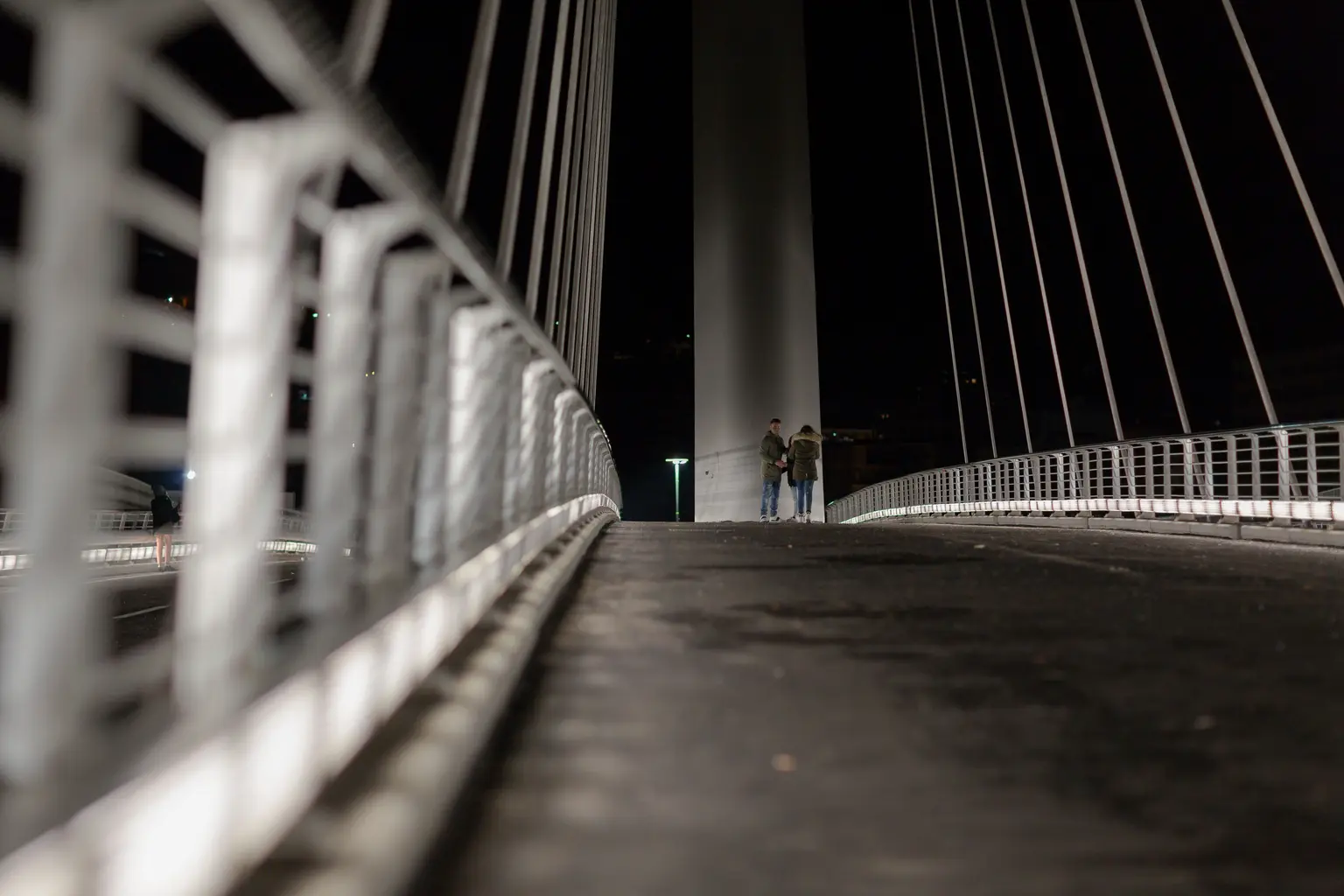 A modern cable-stayed bridge photographed at night, with two people standing near the central pylon. The perspective is low to the ground, highlighting the bridge’s railings and cables.