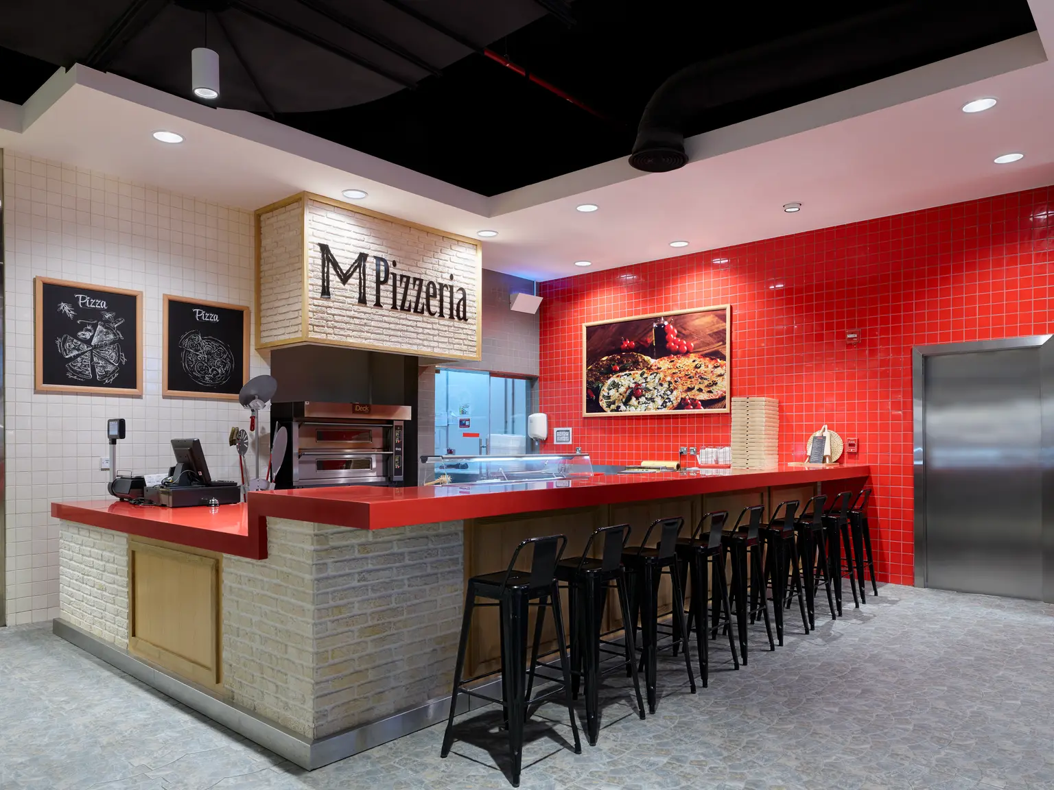 L’interno di una moderna pizzeria presenta una parete con piastrelle rosse, un bancone elegante con sgabelli da bar neri e cucina a vista. Lo spazio è ben illuminato, con un mix di elementi di design industriale e contemporaneo.