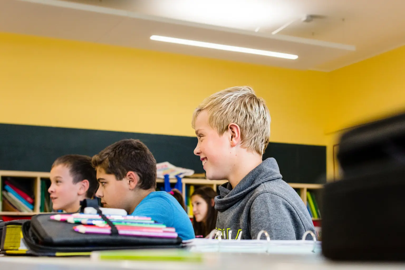 Studenti seduti in un'aula con pareti gialle, una lavagna verde e scaffali pieni di libri e materiali colorati. Il locale è ben illuminato con luci a soffitto e favorisce la concentrazione per l'apprendimento.