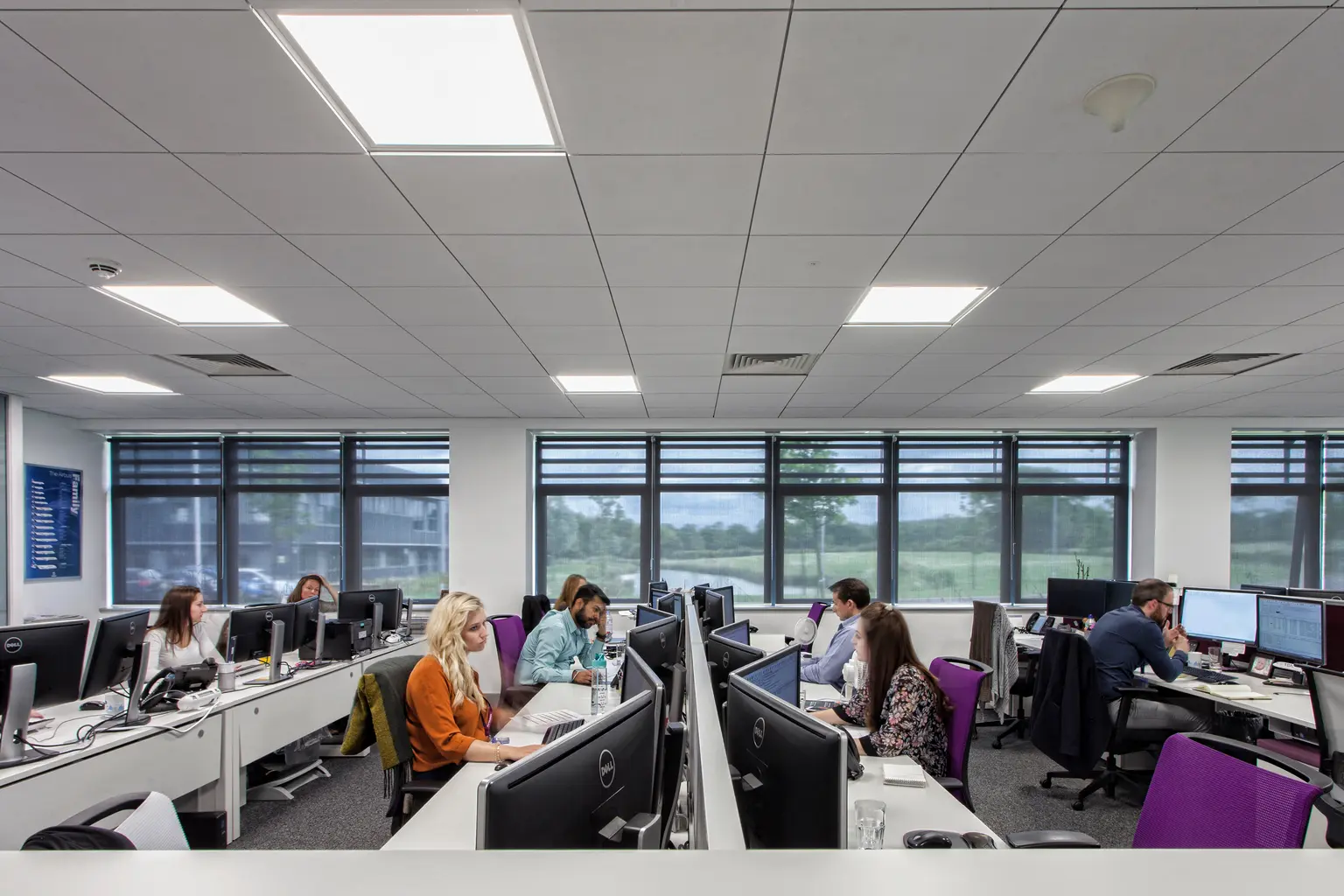 Diverse persone lavorano a scrivanie con computer in un luminoso e moderno ufficio open space con ampie finestre e illuminazione dall'alto. Lo spazio di lavoro ha un'atmosfera collaborativa e una vista sul verde esterno.