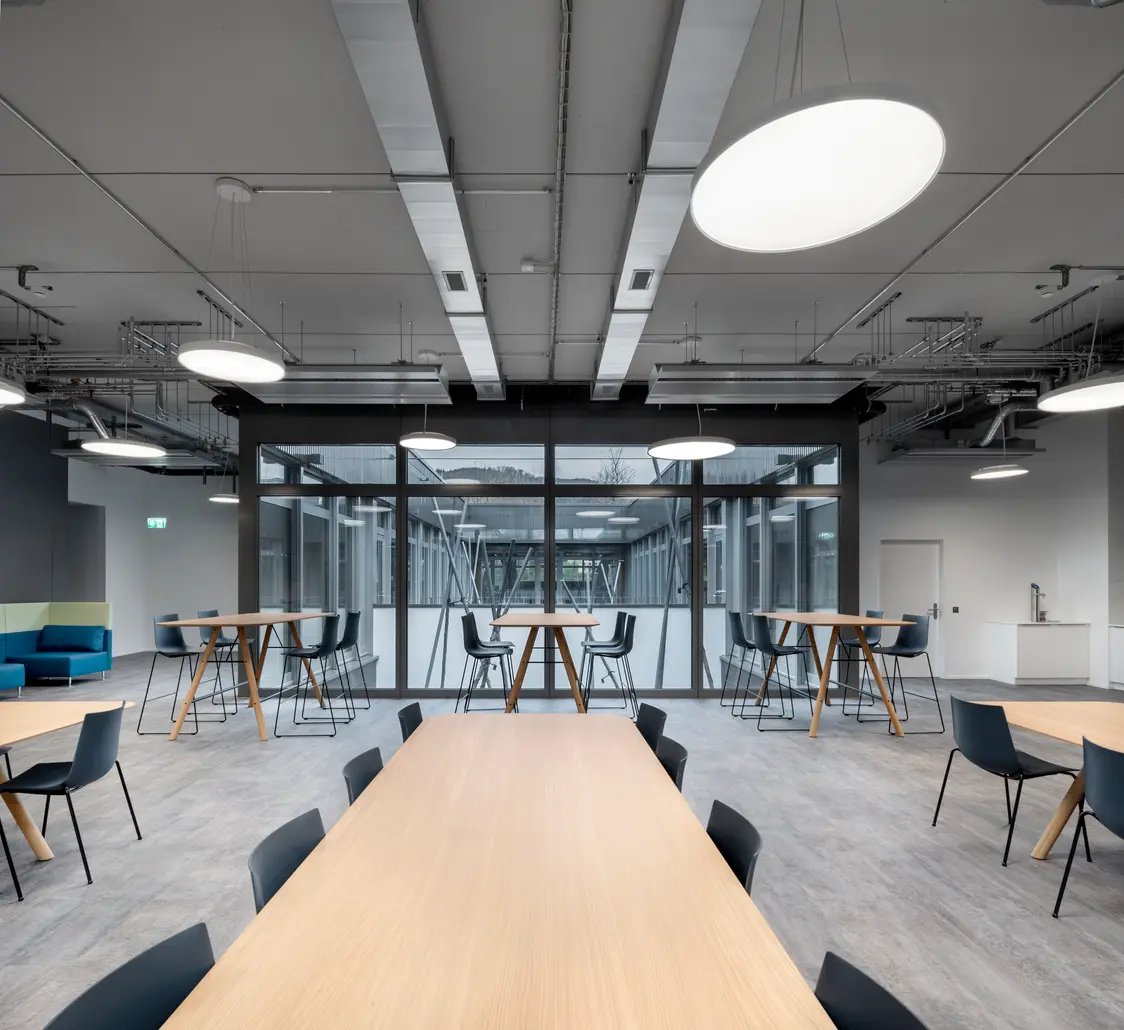 Moderner Aufenthaltsraum mit Holztischen, schwarzen Stühlen und runden LED-Deckenleuchten. Die Glaswand im Hintergrund trennt den Raum und schafft eine helle, offene Atmosphäre.