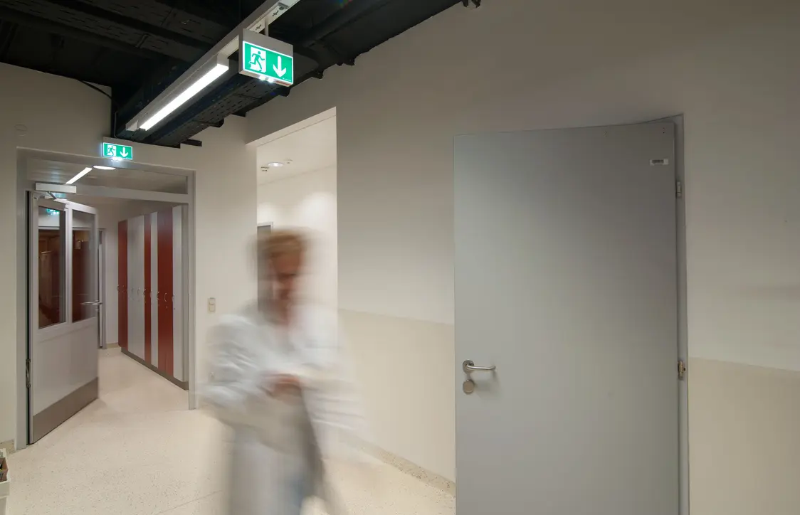 Die CROSSIGN-Leuchte bietet Orientierung in einem Klinikkorridor mit offenen Türen, glatten Wänden und einer verschwommenen Person im Vordergrund.