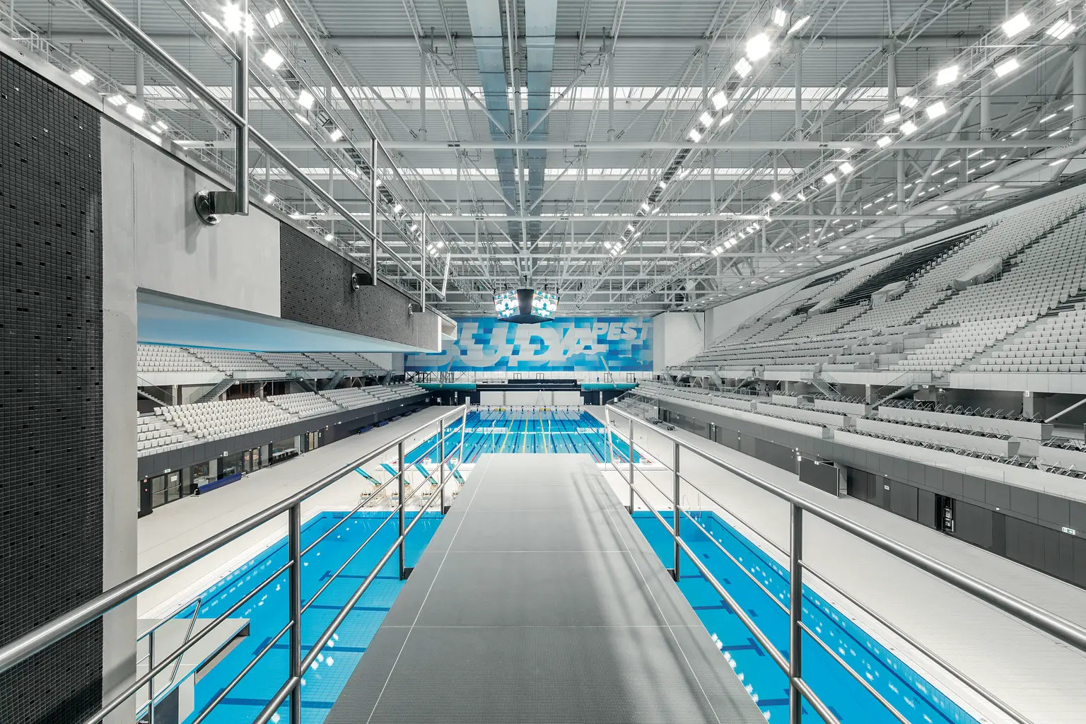Modernes Hallenbad mit mehreren Bahnen, von einem Sprungturm aus betrachtet. Die Halle ist geräumig und hell erleuchtet, mit Reihen von Zuschauerplätzen.