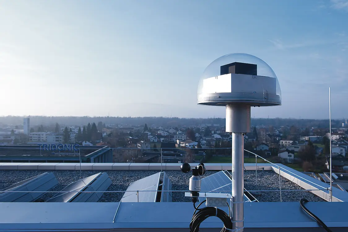 Ein Tageslichtmesskopf mit einem Windgeschwindigkeitsmesser auf einem Dach mit Solarpanelen. Im Hintergrund eine Kleinstadt im Morgengrauen.