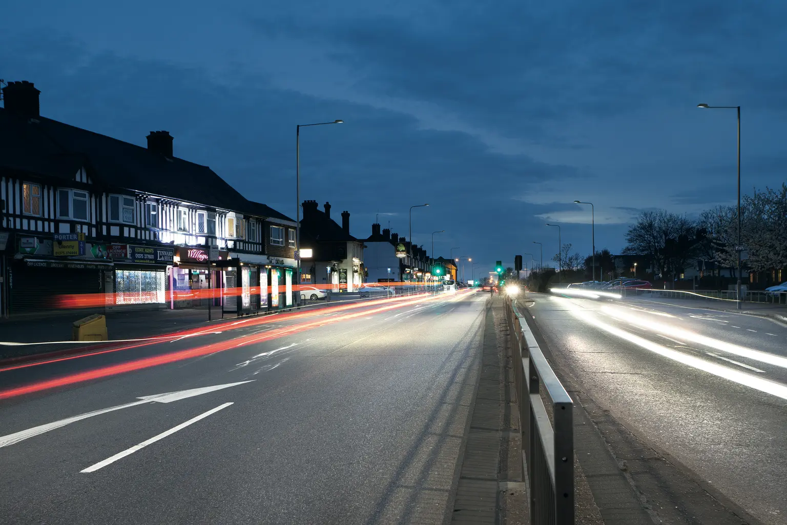 Eine belebte Straße in der Abenddämmerung zeigt Lichtspuren von vorbeifahrenden Autos, mit Gebäuden und Straßenleuchten, die die Straße säumen. Der Himmel ist dunkelblau und die Szene fängt die Bewegung und Energie des Abendverkehrs ein.