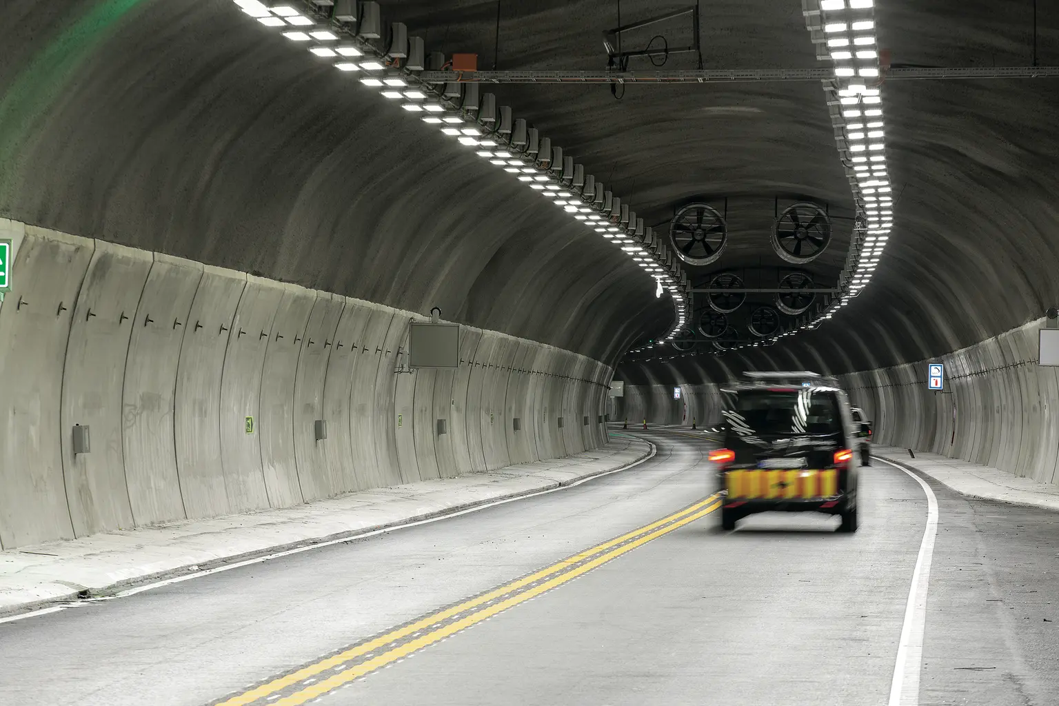 Un tunnel stradale curvo con pareti in cemento liscio e luci LED sul soffitto, luminose e uniformemente distanziate. Un veicolo nero percorre una strada a due corsie segnata da linee gialle e bianche, con grandi ventilatori visibili all'estremità del tunnel.