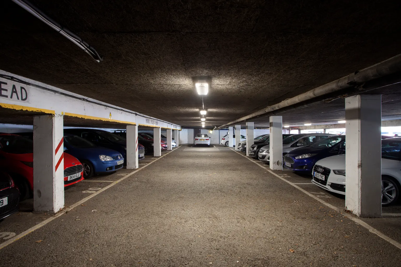 Ein Parkhaus mit Reihen von geparkten Autos auf beiden Seiten und Betonpfeilern, die die niedrige Decke stützen. Der Raum wird mit Leuchtstoffröhren an der Decke beleuchtet.