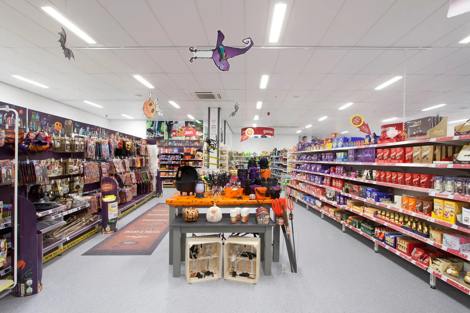 Un corridoio festoso di un supermercato decorato per Halloween, con articoli a tema come zucche, costumi e decorazioni spaventose. Gli scaffali sono riforniti di beni confezionati colorati e il tavolo centrale visualizza prodotti stagionali.