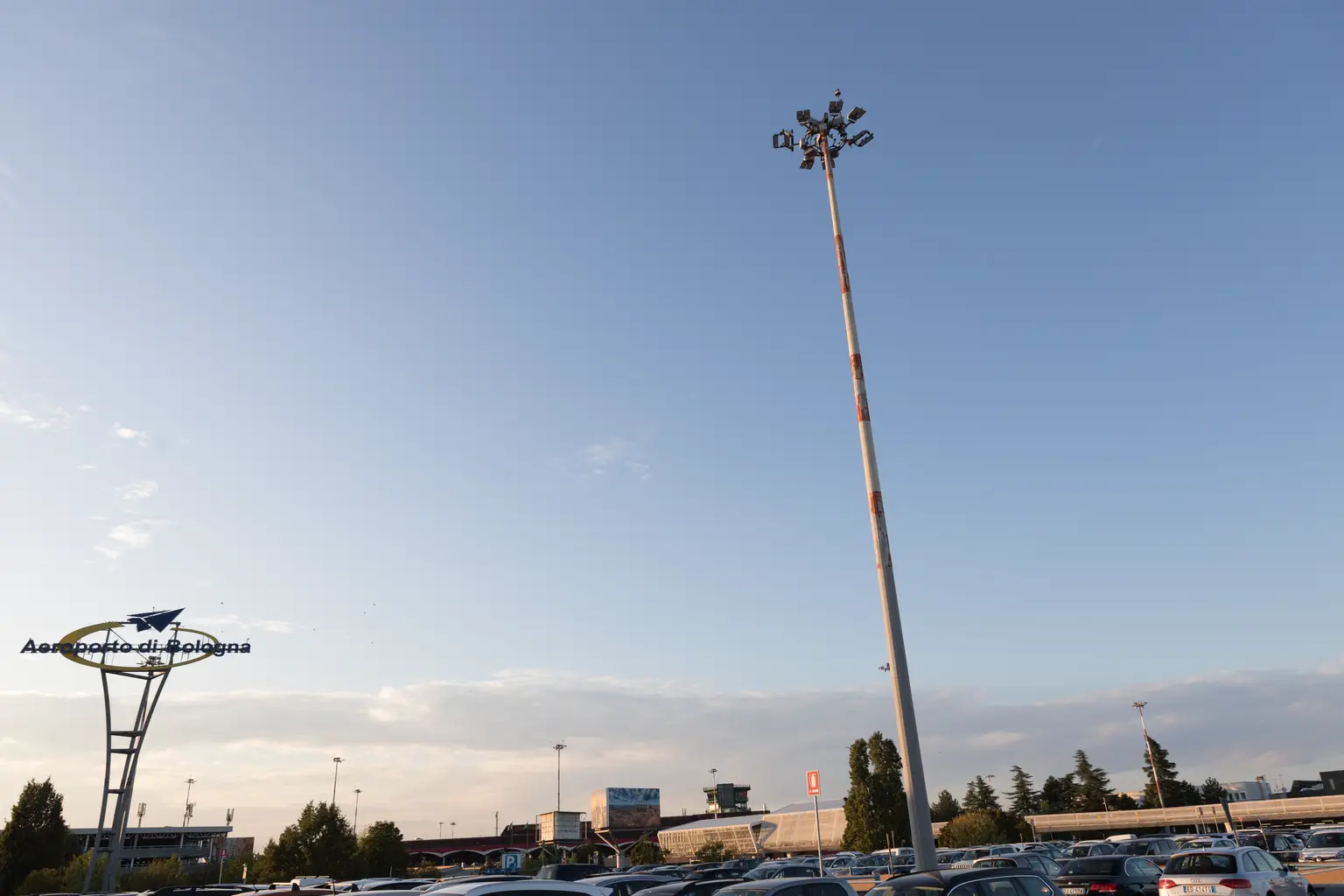 Ampio parcheggio esterno con un alto palo della luce al centro. Diverse auto parcheggiate e un edificio sono visibili sotto un cielo azzurro chiaro.