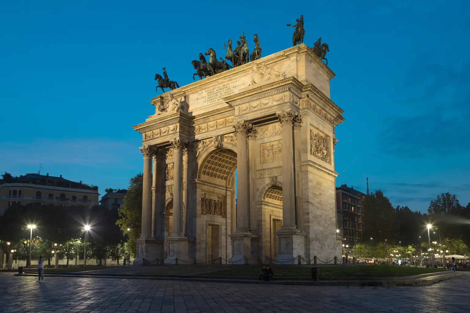 L’Arco della Pace è visibile di lato al crepuscolo, circondato da alberi e luci della città, sotto il cielo azzurro della sera.