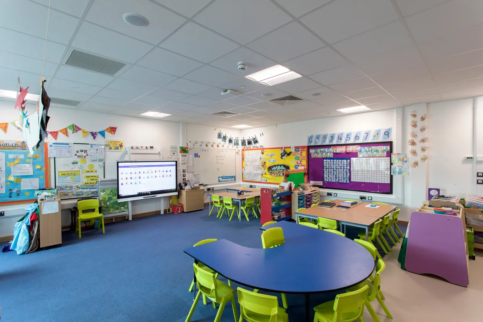Un'aula luminosa e colorata con sedie verdi, un tavolo azzurro e pareti decorate con materiali didattici e disegni degli studenti. Il locale è dotato di lavagna interattiva ed è ben illuminato da luci a soffitto.