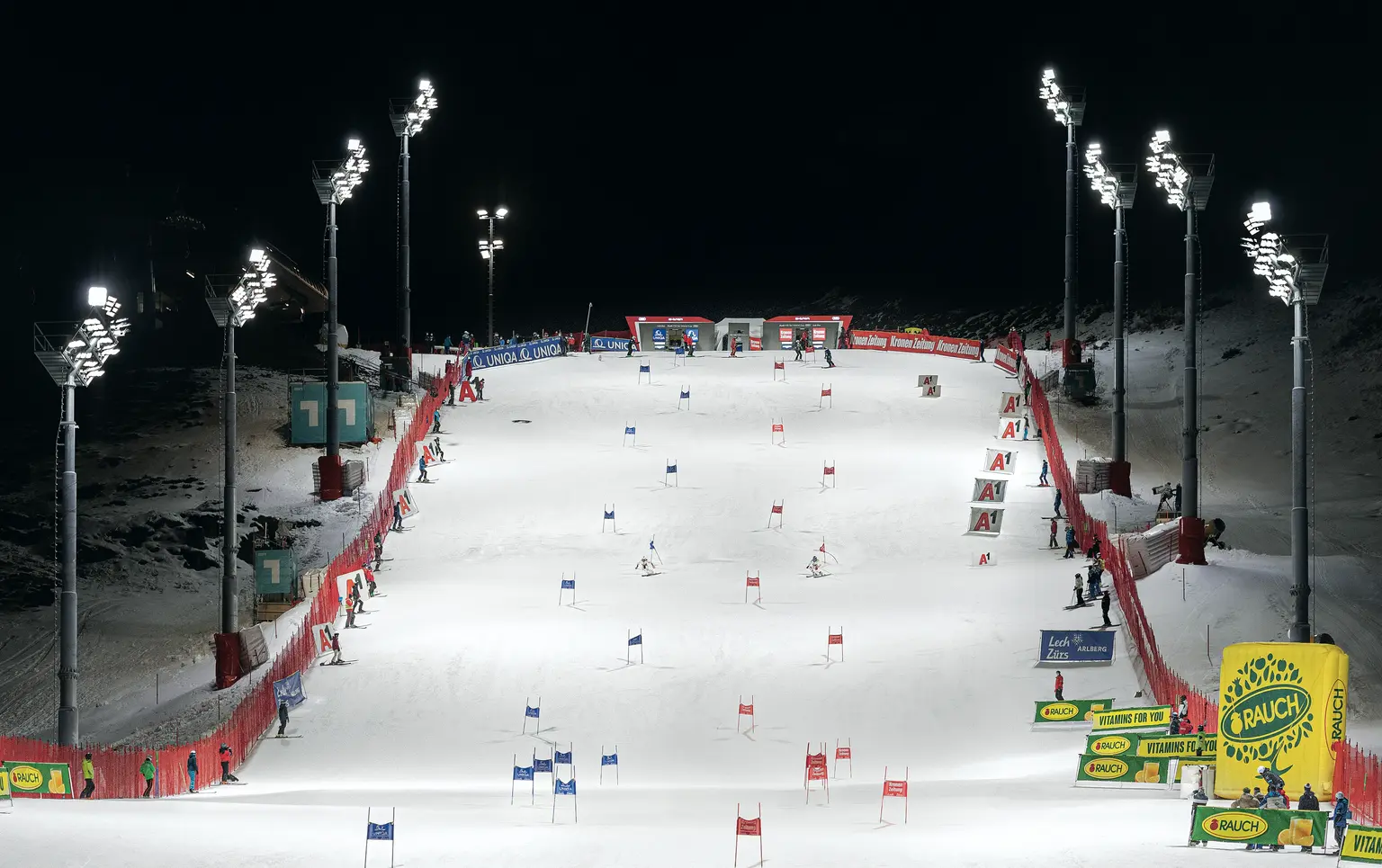 A floodlit ski slope at night with several gates set up for a slalom competition. Floodlight masts and red safety fences line the course, while advertising banners are visible in the finish area.