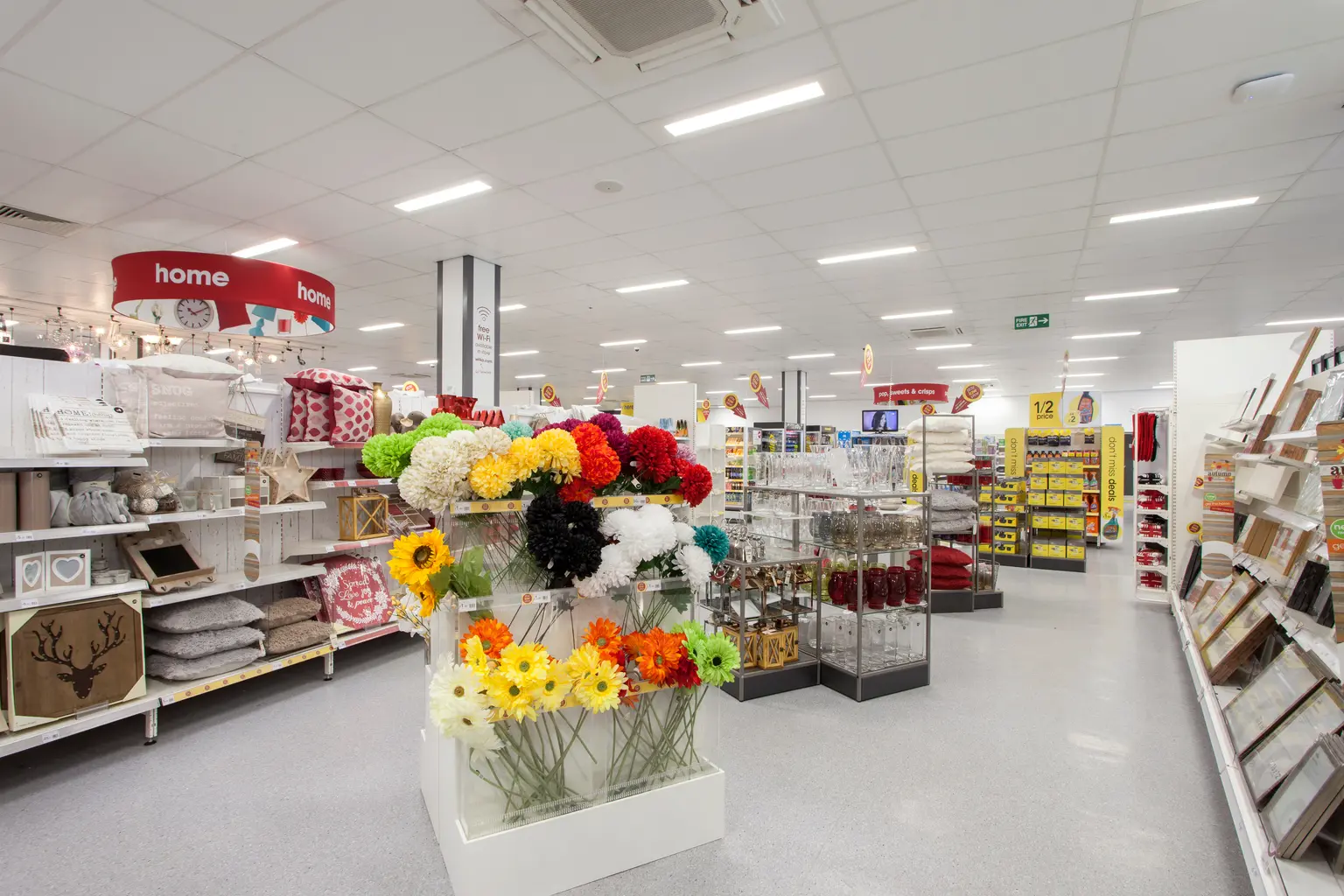 Una sezione di articoli per la casa in un supermercato che espone vivaci fiori artificiali, oggetti decorativi e scaffali ordinatamente disposti. L'area è ben illuminata con pubblicità luminosa etichettata "casa" visibile sullo sfondo.