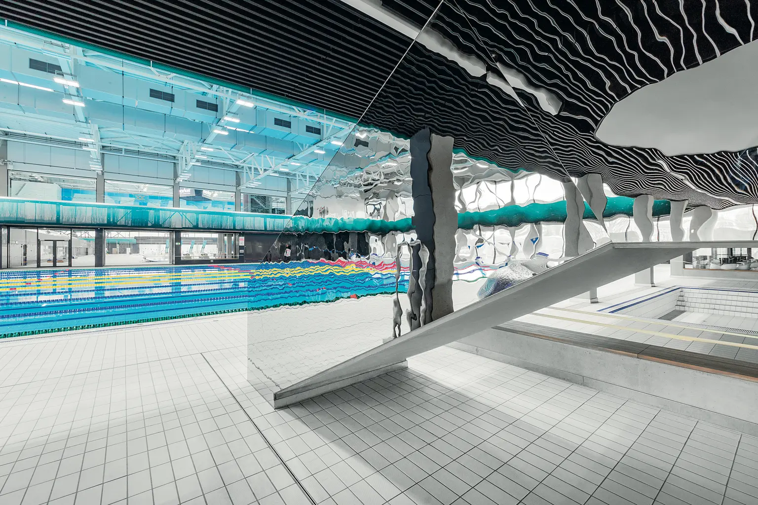 Modernes Hallenbad mit mehreren Bahnen und einem einzigartigen, künstlerischen Deckendesign. Der Bereich ist geräumig und von natürlichem Licht durchflutet, mit einer Rutsche und Sitzgelegenheiten am Pool.