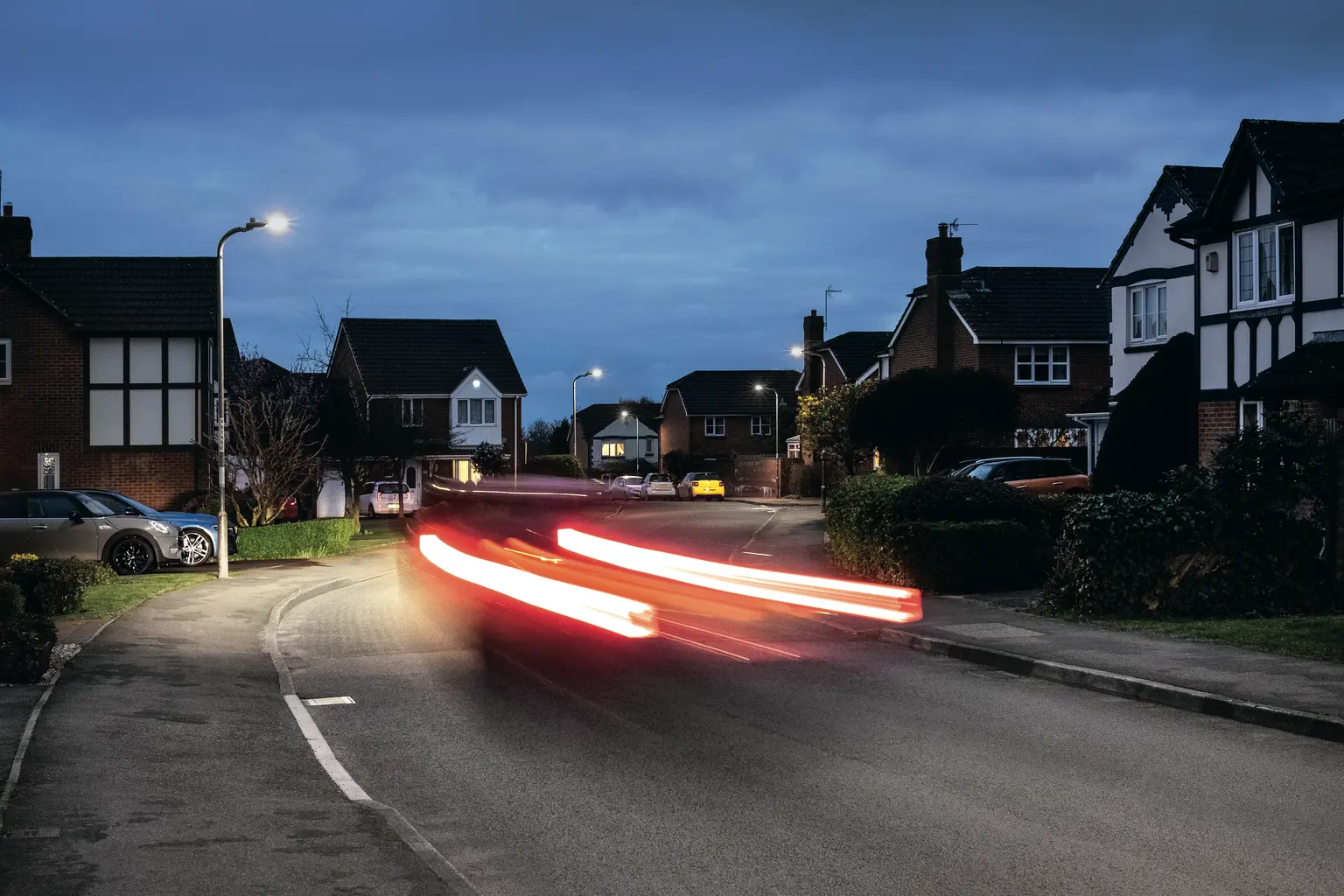 Una tranquilla strada suburbana al crepuscolo con villette e auto parcheggiate su entrambi i lati. Sono visibili le scie di luce rossa di un'auto in transito, e i lampioni illuminano la scena sotto un cielo nuvoloso.
