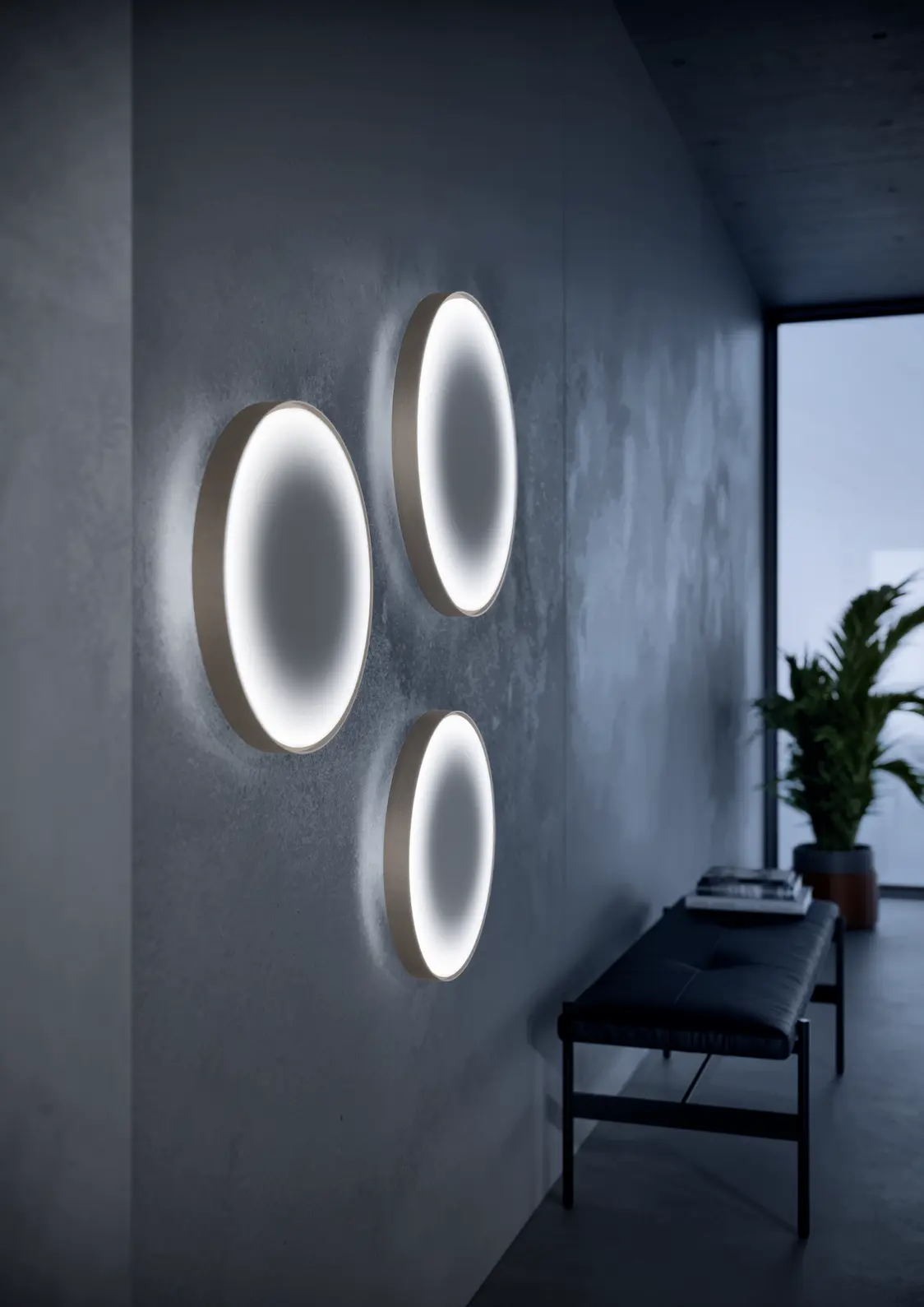Tre apparecchi da parete a LED LANOS in un corridoio moderno creano un'illuminazione morbida e uniforme. Sullo sfondo sono visibili una panchina e una pianta.