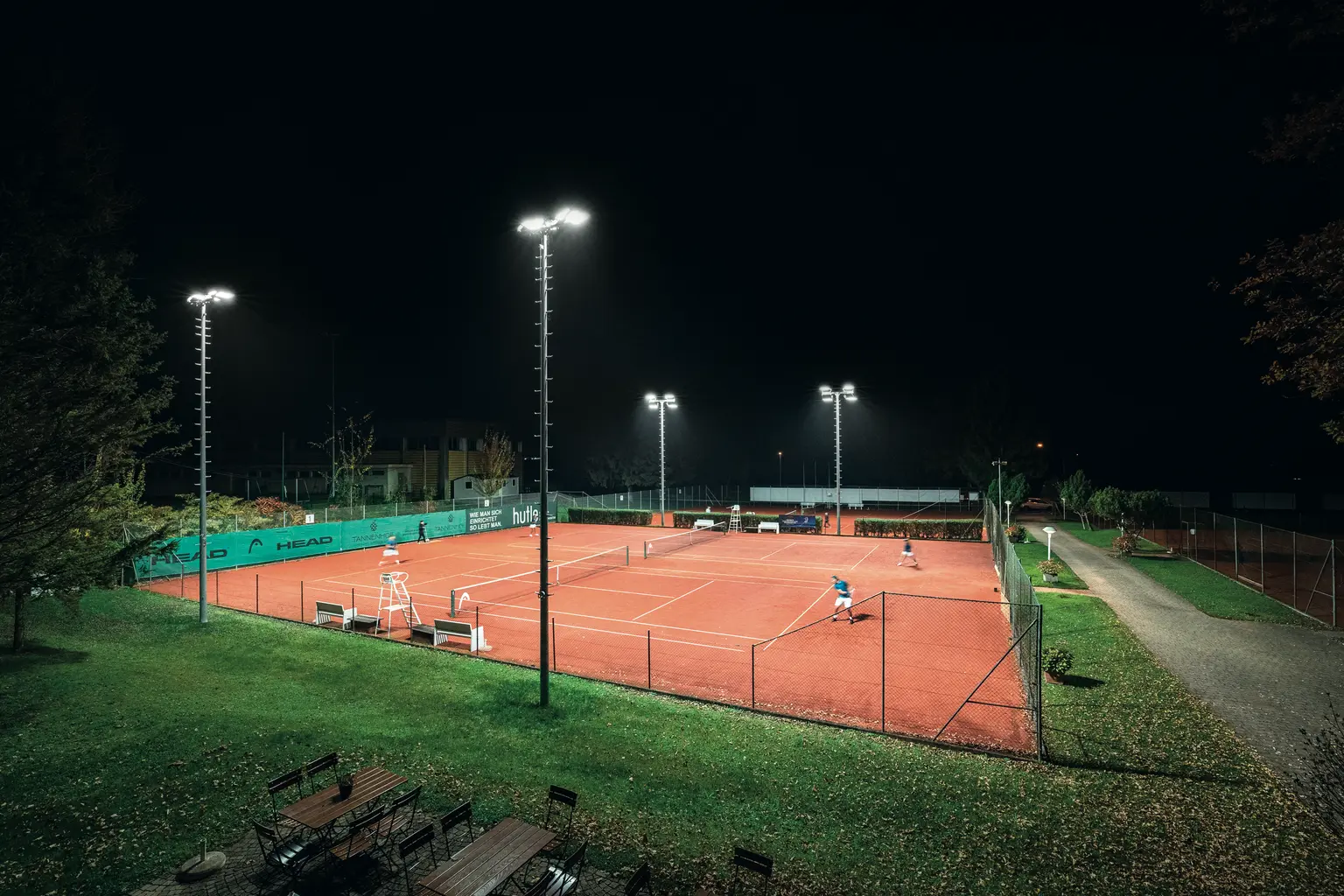 Ein hell beleuchteter Tennisplatz im Freien bei Nacht, umgeben von grünen Zäunen und Bäumen. Die Spieler sind auf dem roten Sandplatz zu sehen, der von hohen Flutlichtern beleuchtet wird.