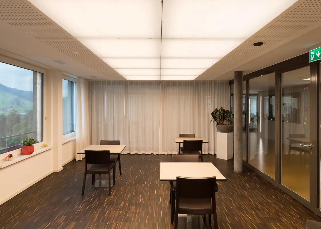 Ein heller, moderner Raum mit Tischen und Stühlen, ideal für Besprechungen oder ruhiges Arbeiten, beleuchtet durch eine innovative Deckenlichtlösung.