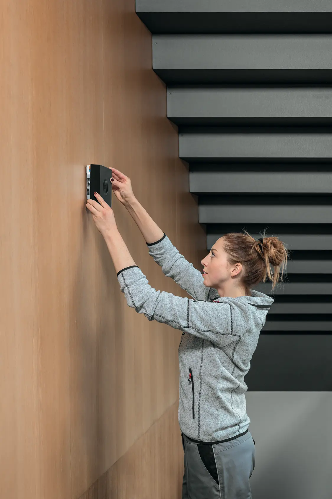 Una donna installa il sistema d'illuminazione RESCLITE PRO su una parte a listelli di legno in una stanza moderna con il soffitto a pannelli scuri e paralleli.