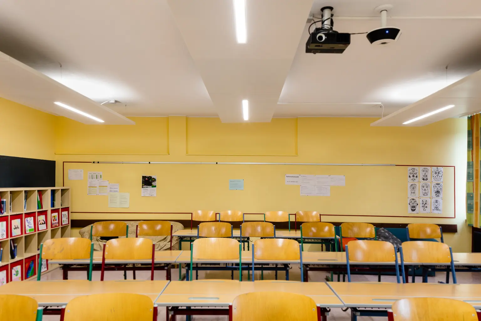 Un'aula luminosa e organizzata con pareti gialle, banchi di legno e sedie abbinate. Il locale è dotato di una lavagna, scaffali per lo stoccaggio e luci montate a soffitto per un'illuminazione ottimale.