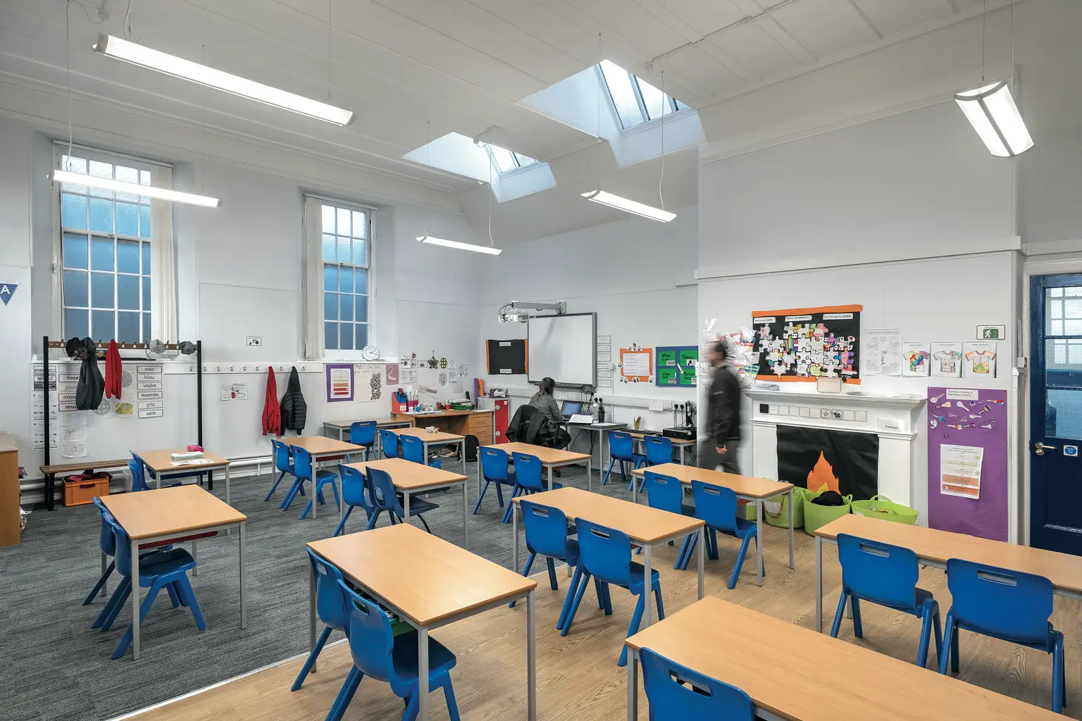 Un'aula luminosa e spaziosa con banchi in legno, sedie blu e ampie finestre che permettono alla luce naturale di entrare. Il locale è dotato di display educativi, una lavagna e luci LED a soffitto per un'illuminazione ottimale.