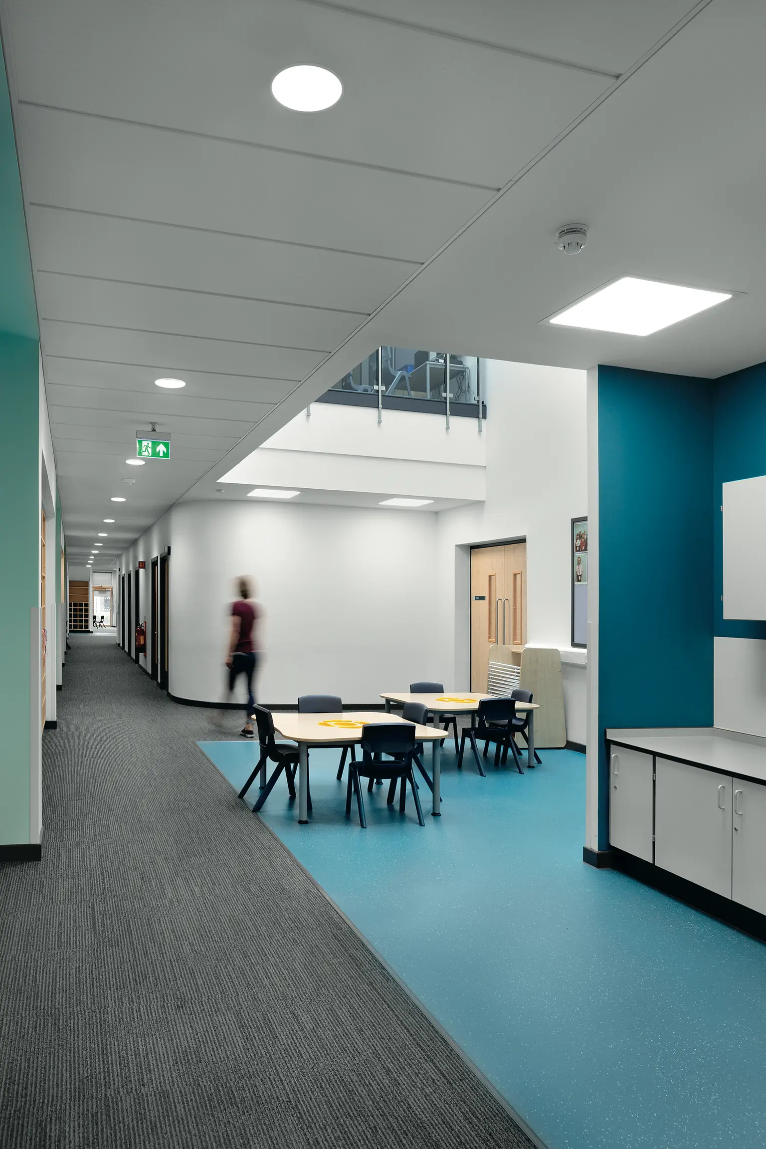 Corridoio scolastico luminoso e moderno con pavimenti in moquette e vinile, pareti bianche e dettagli color foglia di tè. L'ambiente include tavoli rotondi e sedie per attività collaborative, apparecchi a soffitto incassati e una scala che porta al piano superiore.