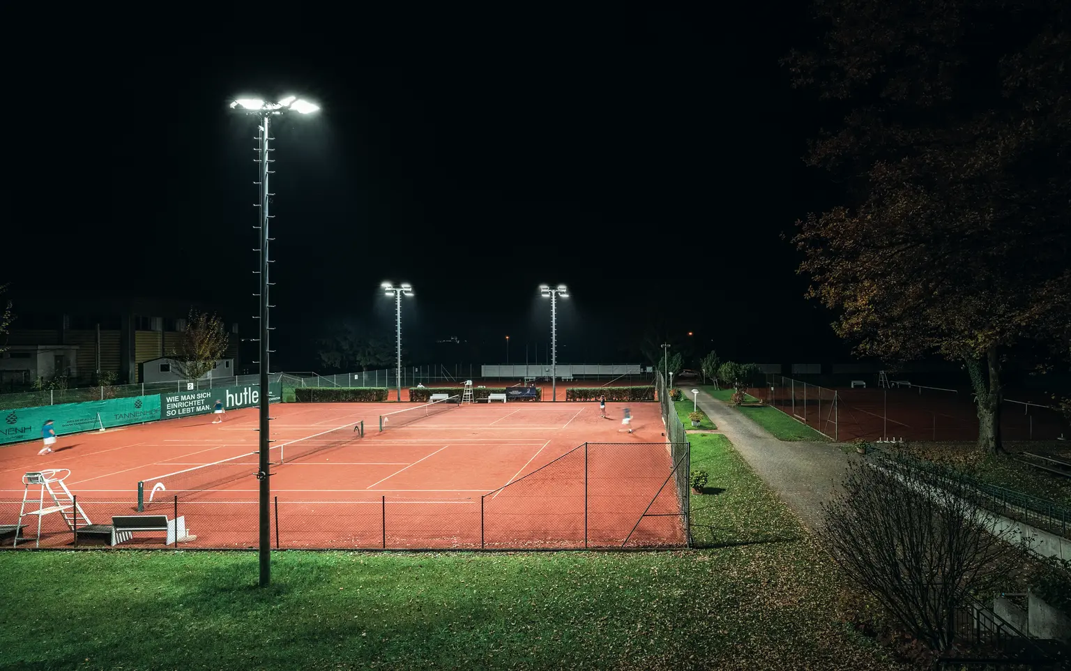 Ein hell beleuchteter Tennisplatz im Freien bei Nacht, umgeben von grünen Zäunen und Bäumen. Die Spieler sind auf dem roten Sandplatz zu sehen, der von hohen Flutlichtern beleuchtet wird.