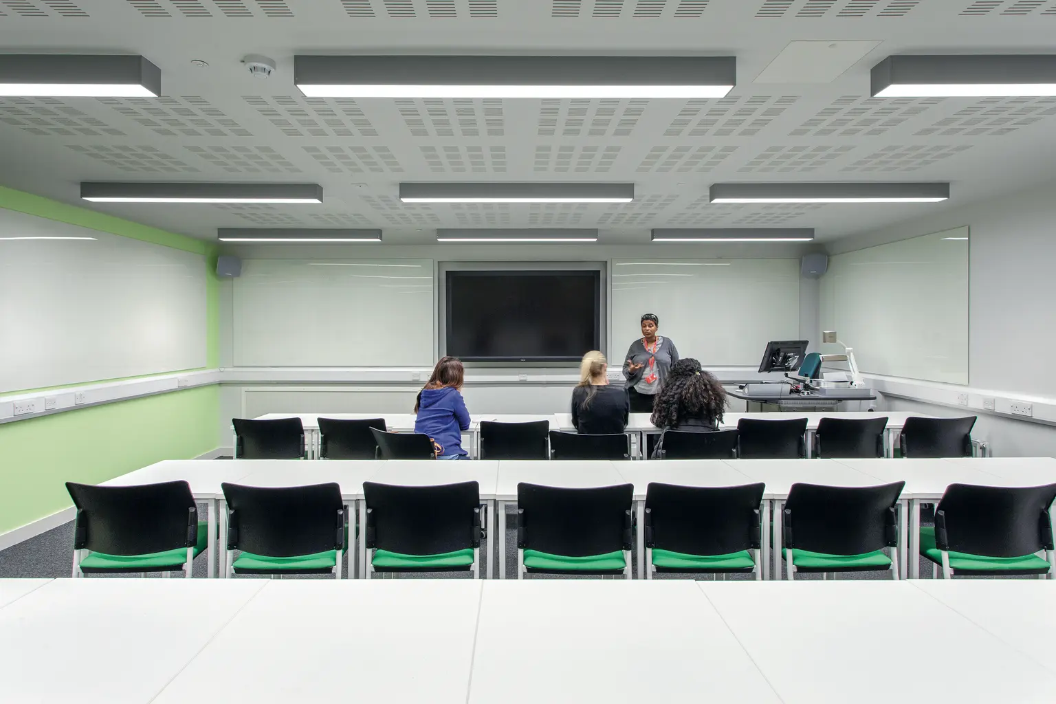 Un'aula moderna con file di scrivanie bianche e sedie nere, dotata di un grande schermo interattivo nella parte anteriore. Il locale è ben illuminato da luci LED montate a soffitto e le pareti presentano elementi decorativi verde chiaro.