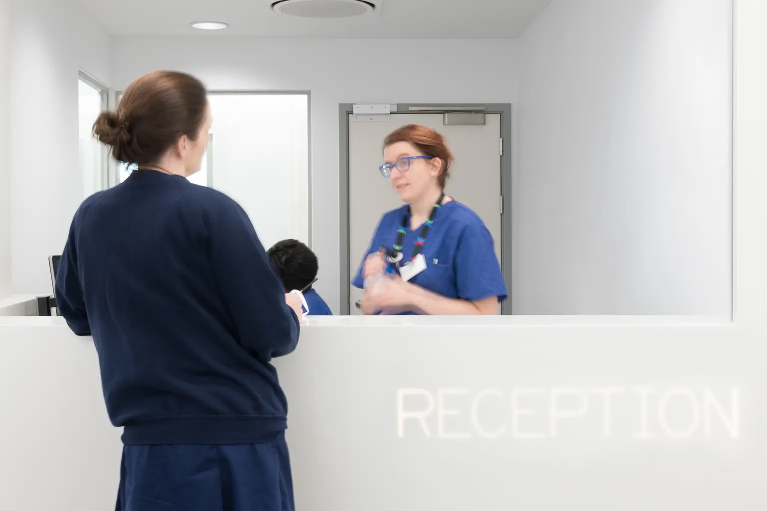Una donna in uniforme azzurro è in piedi davanti a un banco di accettazione bianco e parla con un'altra donna in camice azzurro. Un downlight a LED sullo sfondo illumina l’accettazione.