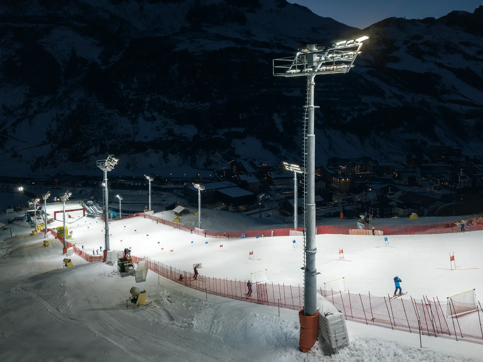 A floodlit ski slope at night with several gates set up for a slalom competition. Floodlight masts and red safety fences line the course, while advertising banners are visible in the finish area.