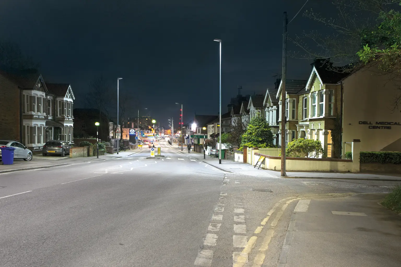 Eine ruhige Wohnstraße bei Nacht wird von Straßenleuchten erleuchtet, mit Häuserreihen auf beiden Seiten. Die Straße wirkt leer, und die Szene ist ruhig und friedlich.