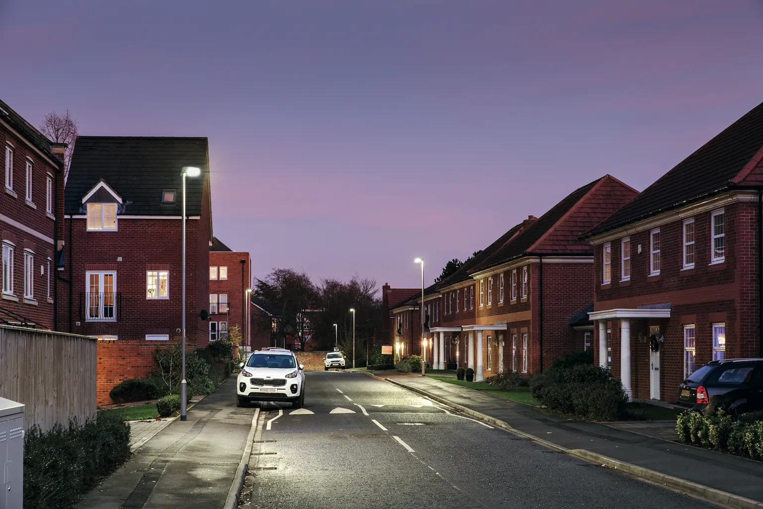 Una tranquilla strada residenziale al crepuscolo è costeggiata da case moderne e illuminata da lampioni. Un'auto bianca è parcheggiata sul lato della strada bagnata, riflettendo la morbida luce della sera.